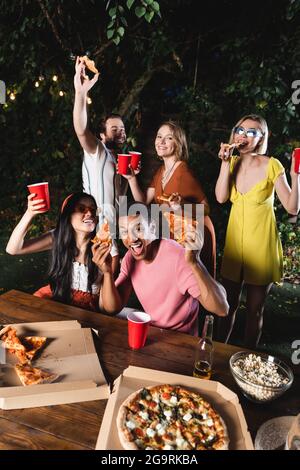 Glückliche multiethnische Freunde halten Pizza und Plastikbecher während der Sommerparty Stockfoto