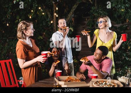 Glückliche multikulturelle Freunde essen Pizza in der Nähe von Plastikbechern und Bier im Freien Stockfoto