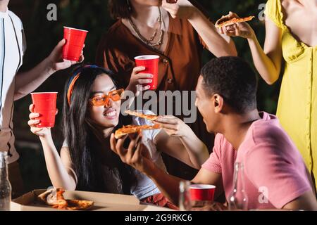 Glückliche multikulturelle Freunde halten Plastikbecher und Pizzastücke im Freien Stockfoto