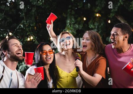 Multiethnische Frauen mit Plastikbechern und Mikrofon singen Karaoke in der Nähe von Freunden Stockfoto