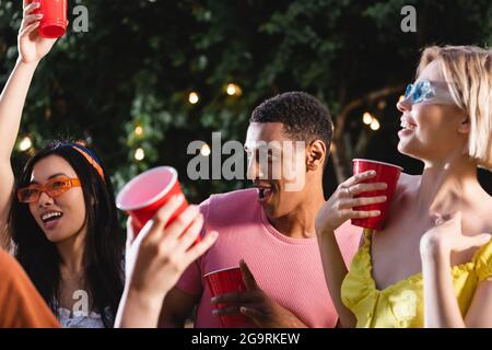 Afroamerikanischer Mann hält Plastikbecher in der Nähe von interracial lächelnden Freunden im Freien Stockfoto