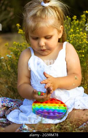 Ein hübsches, kleines blondes Mädchen in einem weißen Kleid spielt im Sommer mit einem Pop auf einer Wiese. Helles Silikon Entwicklung Anti-Stress beliebt Stockfoto