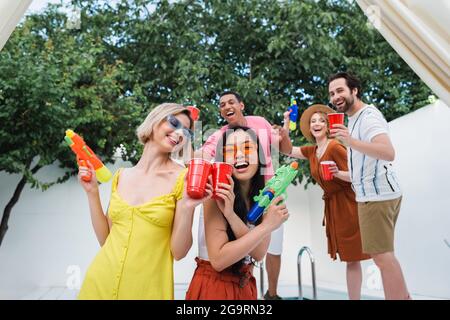 Interracial Frauen mit Wasserpistolen lachen und toasten mit Plastikbechern in der Nähe von glücklichen Freunden Stockfoto