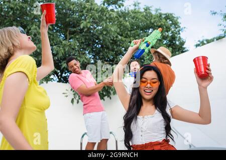 Fröhliche asiatische Frau hält Plastikbecher und Wasserpistole während der Party mit multiethnischen Freunden Stockfoto