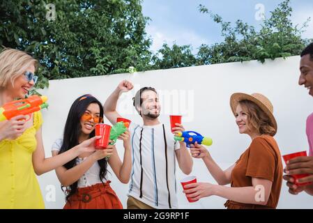 Fröhlicher Mann, der mit Wasserpistolen und Plastikbechern bei multikulturellen Freunden eine Siegesture zeigt Stockfoto