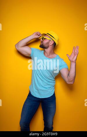 Lebhafter junger bärtiger Rüde in hellblauem Hemd, Jeans und gelbem stylischem Hut, der im Studio steht und gelbe Sonnenbrillen mit der Hand über gelb berührt Stockfoto