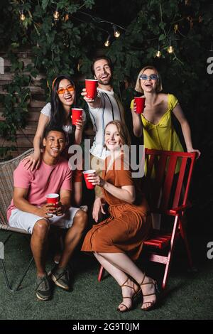 Lächelnde multiethnische Freunde mit Plastikbechern, die während der Party auf die Kamera schauen Stockfoto