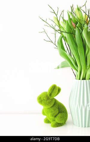 Bouquet von bunten ungeöffneten Tulpen in hellgrüner Keramikvase und Ostergras Hase auf weißem Hintergrund. Urlaubsdekor-Konzept. Stockfoto