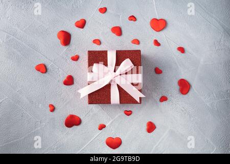 Rote Geschenkbox mit rosa Band und verstreuten roten Herzen auf grauem Hintergrund. Valentinstag- oder Geburtstagskonzept. Stockfoto