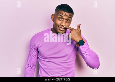 Junger schwarzer Mann mit beiläufigem rosa Pullover, der mit Finger auf Gesicht und Nase zeigt und fröhlich lächelt. Schönheitskonzept Stockfoto