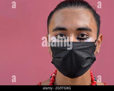 Der junge Latino-Mann mit romantischen braunen Augen und aufwändigem schwarzen Augen-Make-up trägt eine schwarze Corona-Gesichtsmaske zum Einwegnehmen und schaut auf den Betrachter. Stockfoto