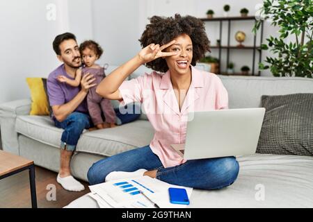 Mutter einer interracial Familie, die zu Hause mit einem Computer-Laptop arbeitet und dabei das Friedenssymbol mit den Fingern über dem Gesicht macht und fröhlich lächelt und den Sieg zeigt Stockfoto