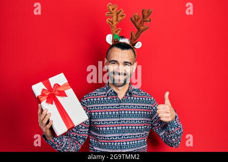 Junger hispanischer Mann trägt Hirsch weihnachtshut mit Geschenk lächelnd glücklich und positiv, Daumen nach oben tun ausgezeichnet und Zustimmung Zeichen Stockfoto