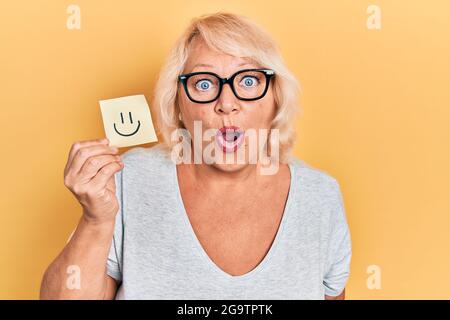 Eine blonde Frau mittleren Alters, die lächelnd erinnert, erschreckte und staunte mit offenem Mund für ein überraschendes, ungläubig gesichtes Gesicht Stockfoto