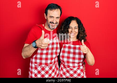 Mittelalterpaar hispanische Frau und Mann tragen Kochschürze tun glückliche Daumen nach oben Geste mit der Hand. Genehmigung Ausdruck Blick auf die Kamera sho Stockfoto