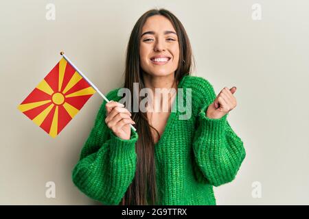 Junge hispanische Mädchen hält mazedonische Flagge schreiend stolz, feiert Sieg und Erfolg sehr aufgeregt mit erhobenem Arm Stockfoto