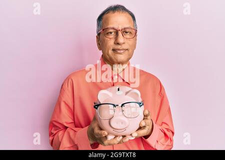 Mittelalter indischer Mann hält Sparschwein mit Brille entspannt mit ernsthaftem Ausdruck im Gesicht. Einfach und natürlich auf die Kamera schauen. Stockfoto