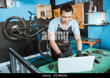 Fahrradmechaniker in Schürze mit Handschuhen und Laptop, um nach Ersatzteilen zu suchen Stockfoto