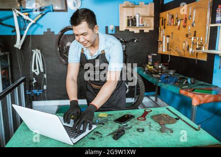 Ein Fahrradmechaniker in einer Schürze, der Handschuhe trägt, wird aufgeregt, während er mit einem Laptop nach Teilen sucht Stockfoto