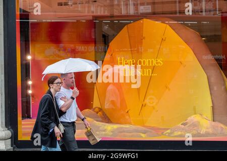 London, Großbritannien. Juli 2021. Ein Paar schützt sich vor Regen unter einem Regenschirm, während sie an einem Schild in einem Schaufenster mit der Aufschrift „Hier kommt die Sonne auf der Oxford Street“ vorbei gehen. Kredit: SOPA Images Limited/Alamy Live Nachrichten Stockfoto