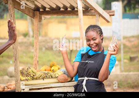 Stock-Foto einer weiblichen afrikanischen Lebensmittelverkäuferin mit Schürze, die an ihrem Stand steht und glücklich Geld, Bargeld oder Naira-Scheine hält Stockfoto