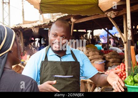 Stock Foto von männlichen afrikanischen Lebensmittelverkäufer oder Geschäftsmann mit Schürze, stand an seinem Stand in einem Markt, bereit, an Kunden zu verkaufen Stockfoto