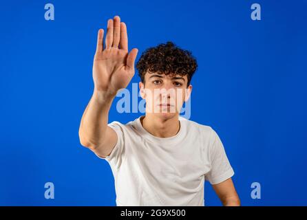 Junger kaukasischer, hübscher Mann, der ein weißes T-Shirt auf blauem Hintergrund trägt, hört ernsthaft auf, mit der Handfläche zu singen. Isoliert auf blauem Studio Stockfoto