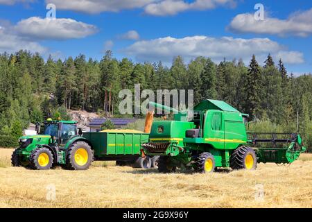 Der Mähdrescher John Deere 9560i WTS entlädt geernteten Weizen auf den landwirtschaftlichen Anhänger hinter dem Traktor John Deere 6155R. Salo, Finnland. 25. Juli 2021 Stockfoto