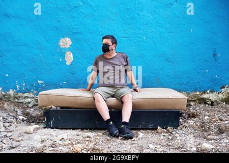 Kaukasischer Mann, der eine medizinische Schutzmaske trägt und auf einem ausrangierten Bett vor einer blauen Wand sitzt. Coronavirus- und Obdachlosenschutzkonzept. Stockfoto