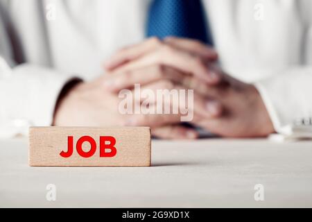 Das Wort Job auf einem Holzblock vor einem Geschäftsmann mit gekreuzten Händen geschrieben. Stellenbewerbung oder Konzept der Beschäftigungsmöglichkeit. Stockfoto