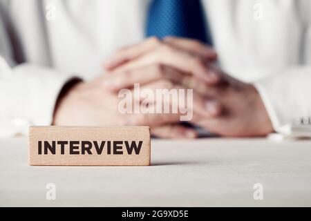 Das Wort Interview geschrieben auf einem Holzblock vor einem Geschäftsmann mit gekreuzten Händen. Konzept für Bewerbungsgespräche. Stockfoto