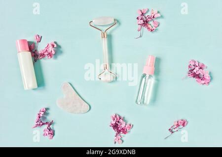 Gua Sha Massagegerät und Walze aus Rosenquarz auf blauem Hintergrund. Gesichtsserum oder Massageöl und Lotion. Rosafarbene Blüten in der Umgebung. Flach liegend, Draufsicht. Stockfoto