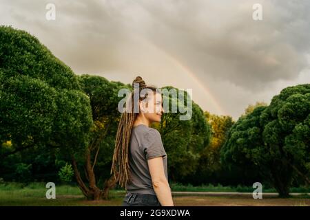 Profilansicht einer lächelnden jungen Frau im Park. Regenbogen auf dem Hintergrund als Symbol alles gut. Speicherplatz kopieren. Stockfoto