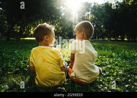 Zwei glückliche Jungen essen Eis und reden auf Gras im öffentlichen Park. Rückansicht. Stockfoto