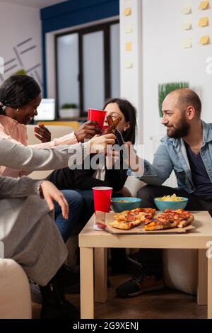Verschiedene Gruppen von Mitarbeitern, die nach der Arbeit auf der Büromeeting-Party Spaß haben. Fröhliche Freunde feuern Flaschen und Tassen Bier an, um die Pause vom Geschäft zu feiern. Multi-ethnische Menschen lächeln Stockfoto