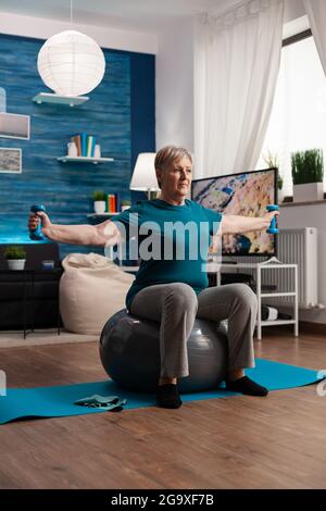 Sportlicher Rentner in Sportswear, der online Aerobic-Workout mit einem Tablet auf schweizer Ball beobachtet und dabei Körperwiderstand trainiert. Ältere Frau führt im Wohnzimmer mit Hanteln eine Armübung durch Stockfoto