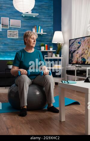 Fokussierte ältere Frau beim Armtraining mit Workout-Hanteln, die auf schweizer Ball im Wohnzimmer sitzen. Rentner in Sportbekleidung, die Online-Aerobic-Training auf Tablet Ausübung Körperwiderstand Stockfoto