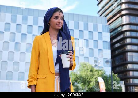 Junge indische Geschäftsfrau in gelber Jacke lächelnd und mit einer Tasse Kaffee im Freien. Stockfoto