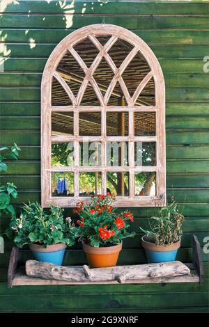 Dekorativer Bogenspiegel an einer grünen Holzwand mit bunten Blumen in den Blumentöpfen. Einrichtung im Landhausstil. Stockfoto