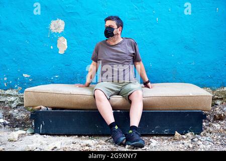 Kaukasischer Mann, der eine medizinische Schutzmaske trägt und auf einem ausrangierten Bett vor einer blauen Wand sitzt. Coronavirus- und Obdachlosenschutzkonzept. Stockfoto