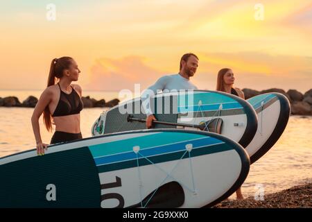 Sommerzeit. Eine Gruppe von Leuten, die mit Brettern am Strand spazieren gehen. Im Hintergrund das Meer und der Sonnenuntergang. Speicherplatz kopieren. Surfen. Stockfoto