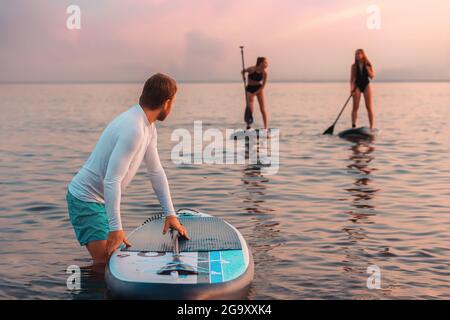 Sommerzeit. Surfer schwimmen an den Surfbrettern am Meer. Mann, der eine Gruppe junger Frauen ansieht. Im Hintergrund das Meer und der Sonnenuntergang. Surfen und rec Stockfoto