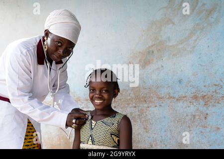 In diesem Bild untersucht eine schwarze Krankenschwester die Lunge und das Herz eines lächelnden, selbstbewussten kleinen afrikanischen Schulmädchen mit einem Stethoskop in einem ländlichen Krankenhaus Stockfoto