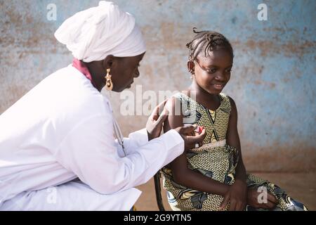 Auf diesem Bild platziert eine schwarze Krankenschwester sanft ein modernes Digitalthermometer unter den Arm eines lächelnden kleinen afrikanischen Schulmädchen mit Fieber Stockfoto