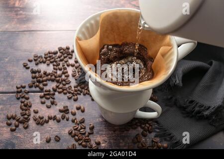 Gebrühten Kaffee, Gießen heißes Wasser auf gemahlenen Kaffee in einem Filter auf einem Becher für ein anregendes Getränk, rustikalen Holztisch mit einigen Bohnen, kopieren Raum, selec Stockfoto