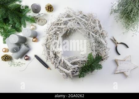 Dekorieren eines Adventskranzes aus weißem Holz mit grünen Feuerästen, blassgoldenen Weihnachtskugeln und grauen Kerzen für ein natürliches Winterarrangement, h Stockfoto