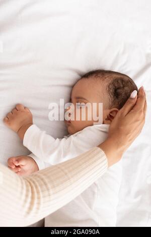 Schwarze Mutter streichelt mit der Hand und schläft den Kopf des Babys im Bett, abgeschnitten Stockfoto