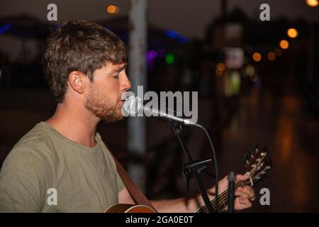 Straßenmusiker singt ein Lied und spielt abends Gitarre. Konzentrierter Sänger, der mit einem Mikrofon und einem Instrument auftrat Stockfoto