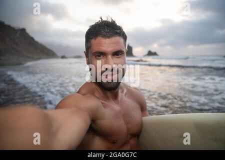 Surfer macht ein Selfie am Strand während des Sonnenuntergangs, er hält sein Surfbrett und lächelt. Junger attraktiver Mann am Strand - fröhliche Person beim Üben Stockfoto