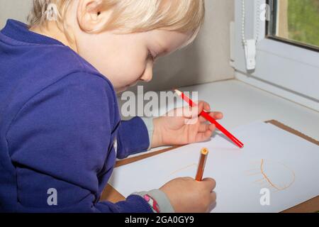 Ein kleiner, hellhaariger Junge mit Bleistiften in den Händen zeichnet auf ein weißes Blatt auf der Fensterbank. Frühkindliche Entwicklung. Stockfoto